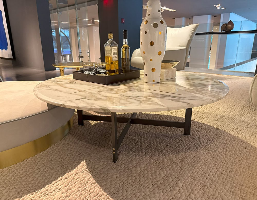 Bax Place Coffee Table with a round marble top featuring light veining and dark metal legs. The surface holds a white vase and a tray with bottles, blending into a relaxed lounge style setting with soft textures and contemporary seating.