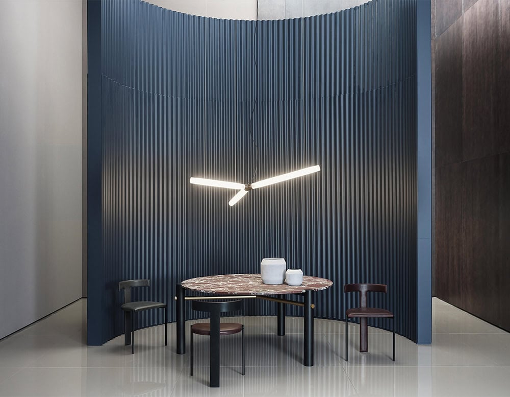 Therna Ceiling Light floats above with sharp linear precision, casting a focused glow over the sculptural setting. A round marble table anchors the space, surrounded by sleek black chairs, all framed by a dramatic, textured blue backdrop.