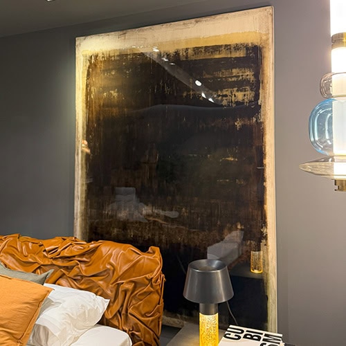 The Dra Sintensi Art is placed above a bed with a caramel leather headboard. Its textured dark composition complements the space, accompanied by a black bedside lamp with a yellow base and stacked design books.