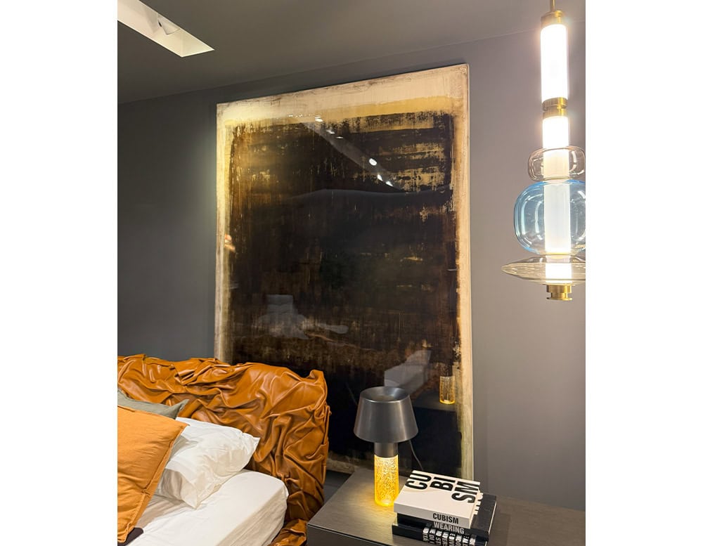 The Dra Sintensi Art hangs in a bedroom setup, its dark textured canvas contrasting with a caramel leather headboard. A bedside table with a yellow-glass lamp and books completes the intimate atmosphere.