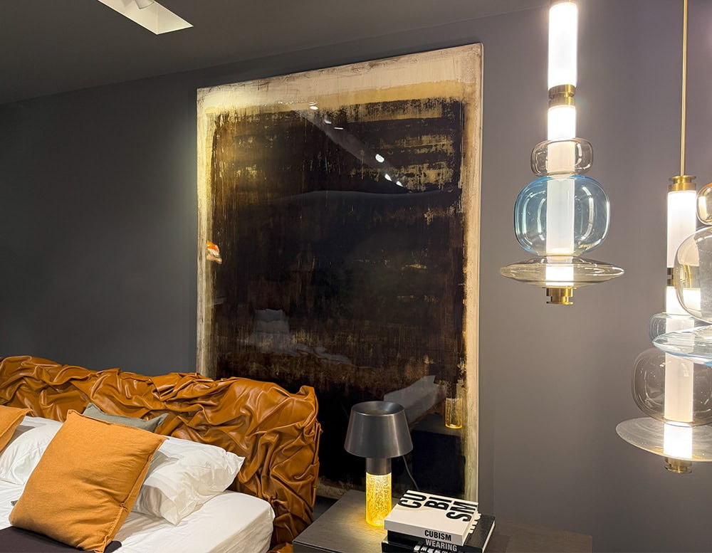 The Dra Sintensi Art is displayed in a bedroom scene, its rich, textured surface positioned above a caramel leather headboard. A stack of books, a yellow-glass lamp, and pendant lights complete the ambient setting.