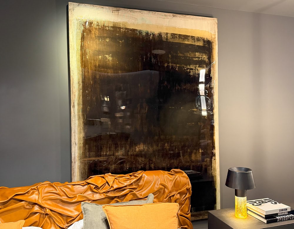 The Dra Sintensi Art is shown mounted on a wall behind a caramel leather sofa. Its dark, layered texture contrasts with the room's soft lighting, while nearby books and a yellow-glass lamp complete the cozy interior scene.