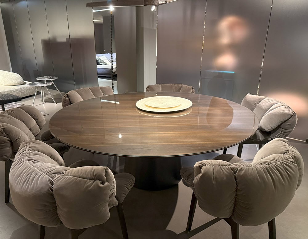 The Emmemobili UFO Dining Table is shown with a round wood veneer top and central pedestal base. It is surrounded by plush tufted chairs in soft grey fabric, creating a cozy and contemporary dining arrangement.