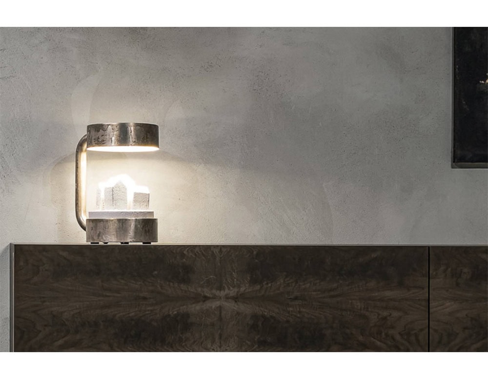 Compaud Table Lamp features a sculptural design with a thick brushed-metal arch and soft white glow that highlights a geometric crystal formation. It blends functional lighting with artistic character and suits modern or industrial interiors.