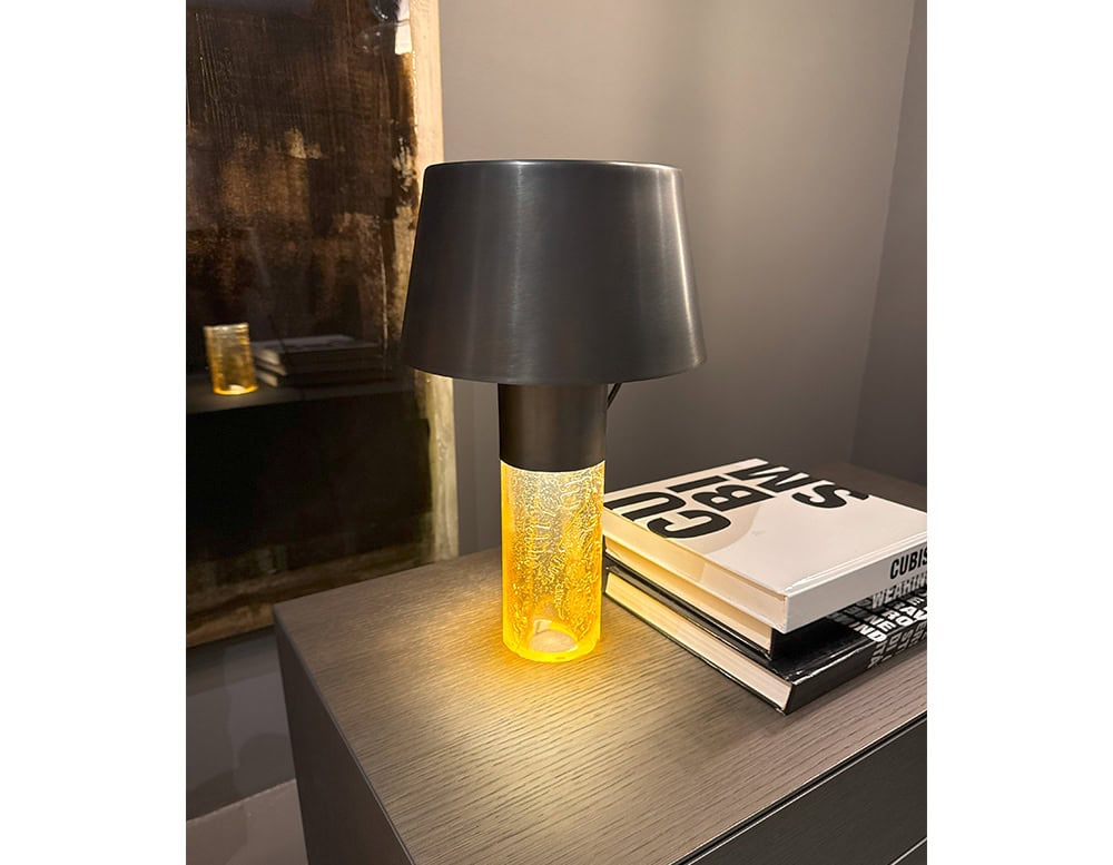 Ghost Table Lamp D25 pairs a dark aluminum shade with an amber glass base that emits a warm, glowing light. The sculptural shape and unique material contrast make it a standout accessory for contemporary side tables or bedroom settings.