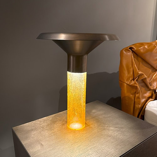 Ghost Table Lamp D38 casts a golden glow through its bubbled amber glass stem, crowned by a dark, flared aluminum top. This unique lamp merges sculptural elegance with soft ambient lighting, ideal for sophisticated interior spaces.