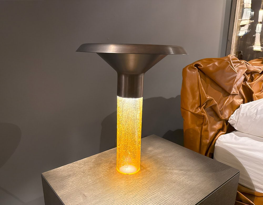 Ghost Table Lamp D38 features a glowing cylindrical base that radiates with a warm, amber hue. Its dark metallic top adds contrast, creating a balanced design that merges artistic lighting with functional style.