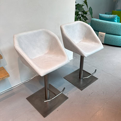 The Hen Hexagon Stools are presented in white upholstery with a sleek, textured finish. Each stool features a brushed steel pedestal and footrest, positioned beside wood shelving and a turquoise modular sofa in a modern lounge setting.