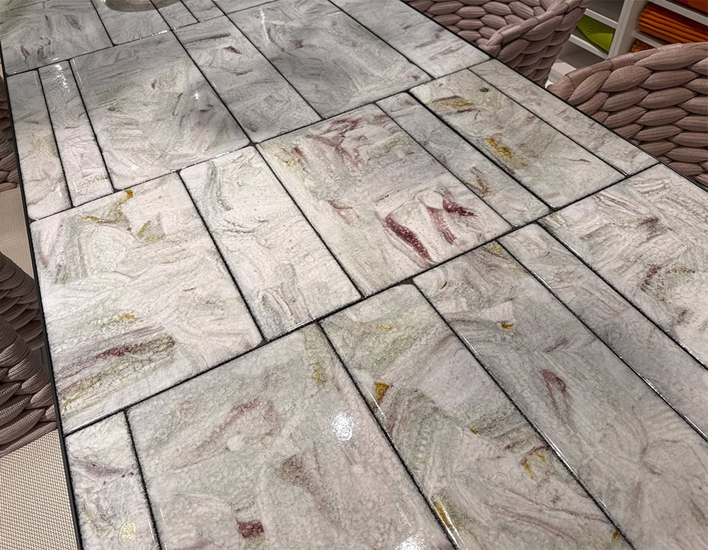 PL Sciara Outdoor Dining Table Gray features a close-up of its glazed ceramic tile top with swirling grays, reds, and yellows. The handcrafted finish gives each tile a unique texture and rich visual character.