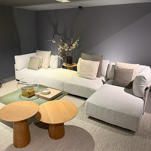 The Tri Nodi Grouping sectional is styled with soft neutral cushions, paired with two round wood side tables and a square green coffee table. A delicate floral arrangement adds an organic accent to this contemporary lounge setup.