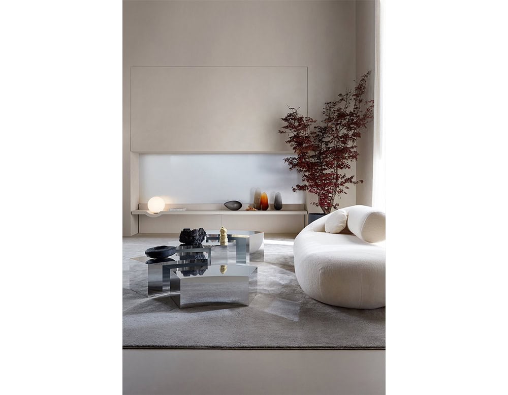 The Evolis Sofa features a soft, rounded form with textured upholstery, perfectly complementing the room’s minimal decor and reflective coffee tables.