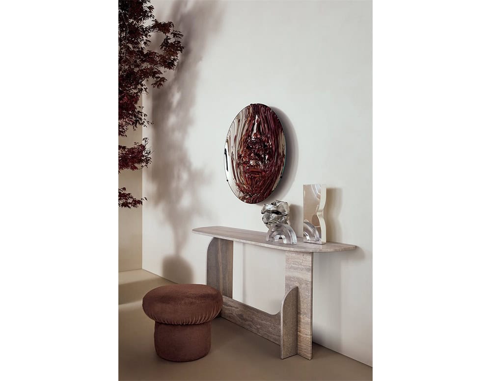 The GR Selce Console Table stands against a neutral wall, complemented by sculptural décor and a reflective art piece above. Its travertine surface and architectural form add a refined, natural texture to the serene interior setting.