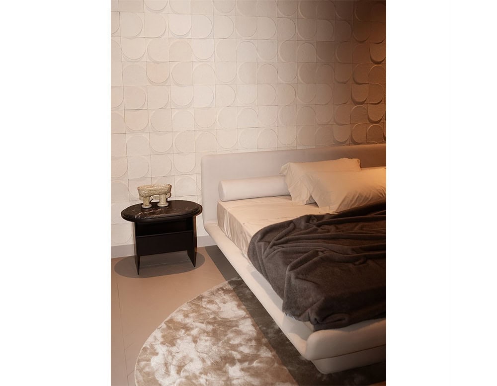 The GR Sensei Night Table stands beside the bed with a dark marble top and black oak base. The cozy bedroom setting features warm lighting, a textured cream wall, and a soft beige and taupe rug.