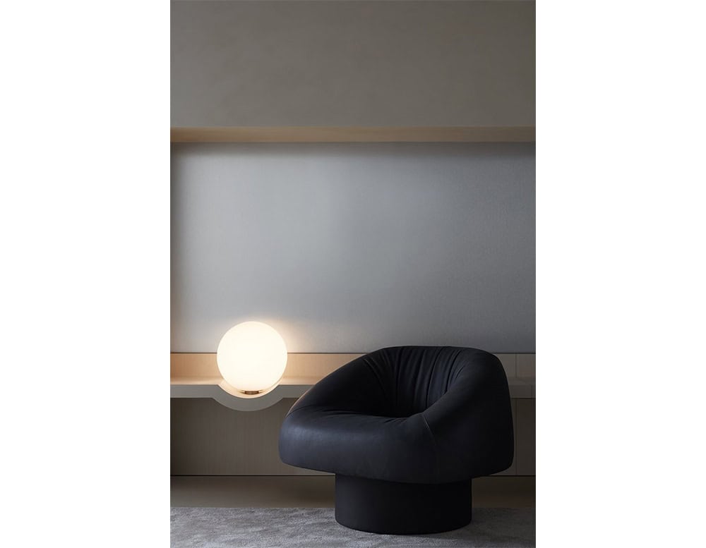 The GR Stresa Console Table features a smooth neutral surface and integrated globe light. It anchors a cozy corner with soft grey walls, warm lighting, and a deep charcoal lounge chair resting on a plush carpet.