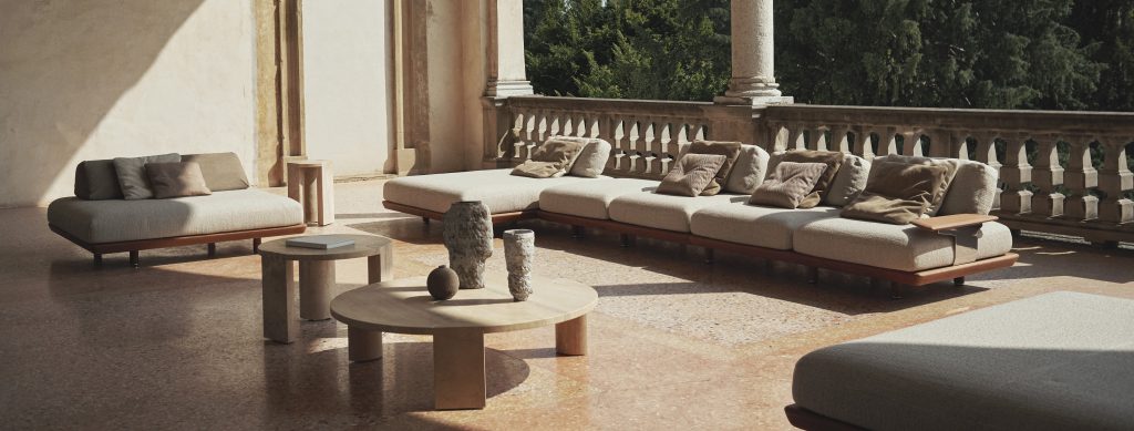 An elegant outdoor terrace featuring modular sofas with soft neutral cushions, complemented by round wooden tables and sculptural decor, set against a sunlit stone backdrop and lush green views.