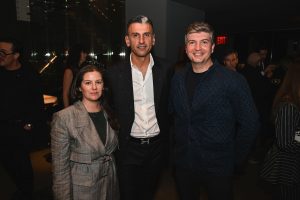 Three guests gather for a photo during the evening event, dressed in polished, modern attire and sharing warm smiles as the lively atmosphere continues around them.