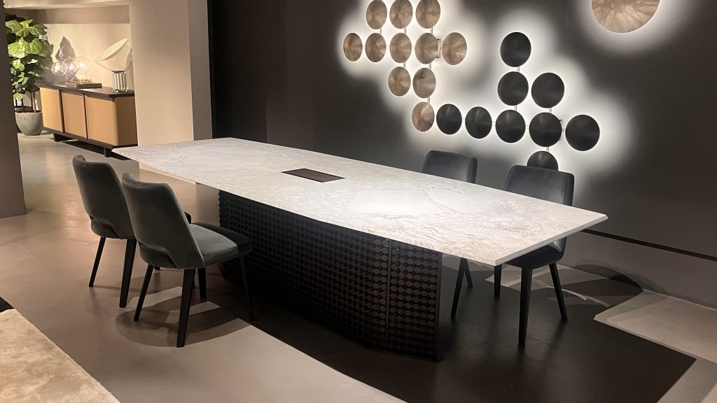 S Penny dining table with a light marble top featuring subtle gray veining, paired with a sculptural dark wood base. Shown with deep charcoal upholstered chairs, the palette blends soft stone tones with rich wood and dark accents.
