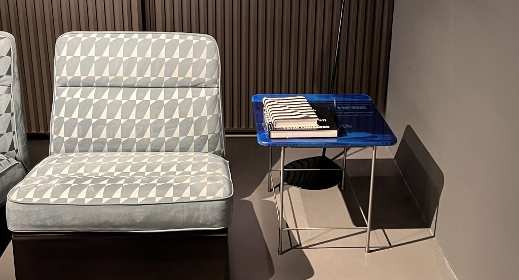 This small accent table features a rich blue glass top with layered tonal variations and subtle graphic details. The glossy surface is supported by a slim silver metal frame, adding a modern touch with a bold pop of color.