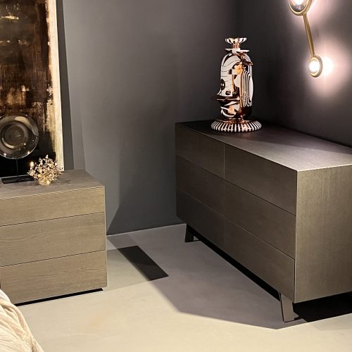 Refined bedroom vignette with coordinated nightstand and dresser in warm taupe wood. Soft gray walls, neutral bedding, and subtle brass lighting create a calm, modern atmosphere with elegant contrast and understated texture.