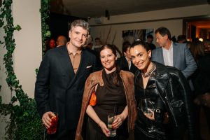 Three guests pose together at a stylish cocktail event, holding drinks and dressed in chic evening attire, while other attendees mingle in the warmly lit background.