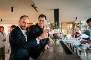 Two guests raise glasses of white wine in a celebratory toast at a bright, modern bar area during a stylish daytime social event, with bartenders and attendees visible in the background.