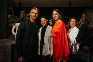 Three guests pose together at an elegant evening event, with one woman wearing a striking bright red dress that stands out against the dark, modern interior and sophisticated cocktail attire.