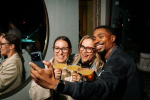 Three friends smile while taking a selfie together at a lively evening gathering, each holding a yellow cocktail in a chic, softly lit indoor setting.