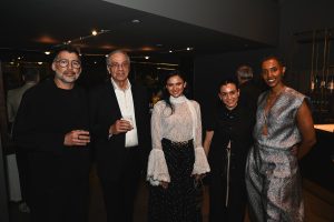 Five guests pose together at an elegant nighttime event, dressed in a mix of sophisticated black, white, and metallic outfits inside a modern showroom with dim ambient lighting.
