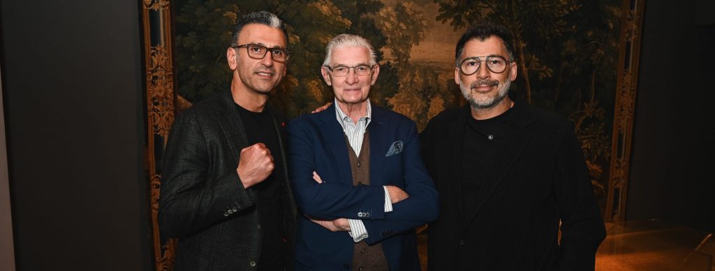 Three men pose together at a sophisticated evening event, standing in front of an ornate tapestry backdrop while dressed in elegant dark tailoring and contemporary formalwear.