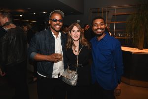Three guests smile for a photo at a stylish evening gathering, dressed in modern cocktail attire inside a contemporary showroom with warm ambient lighting.