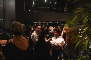 Guests mingle in a lively, modern showroom setting, enjoying conversation, cocktails, and an intimate evening atmosphere under dramatic lighting.