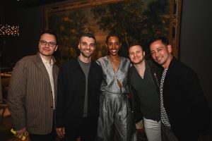 A stylish group gathers for an evening celebration, posing against a dramatic mural backdrop with contemporary fashion and a warm social atmosphere.