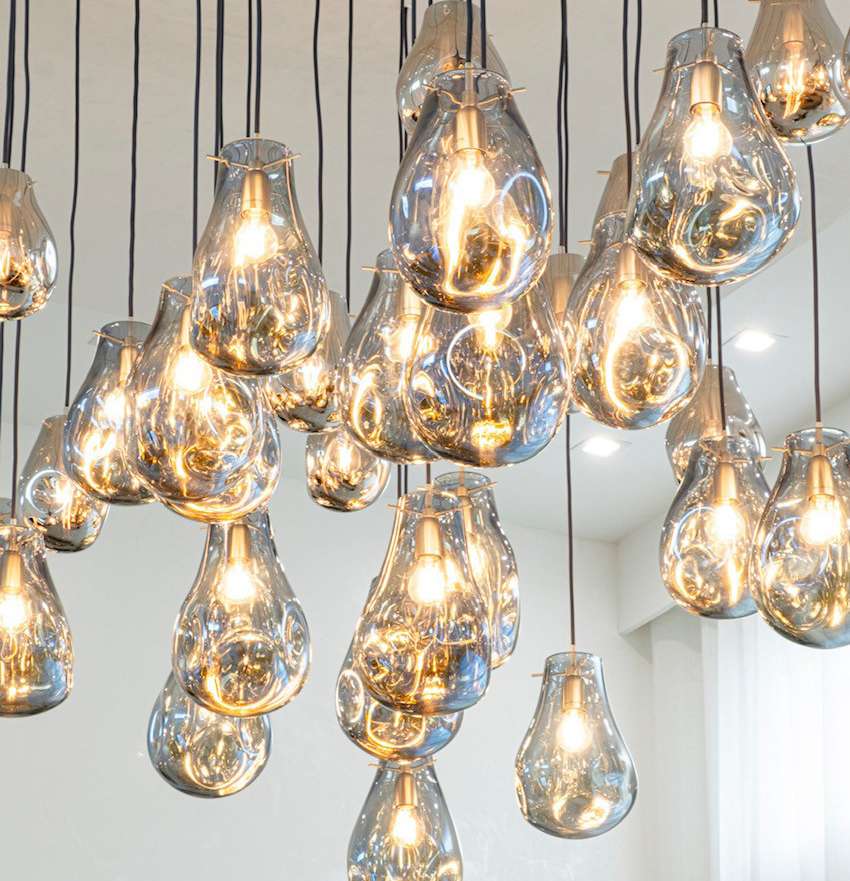 A cluster of hand blown glass pendant lights with soft rounded silhouettes hangs at varying heights. Their transparent smoky finish and warm illumination create layered reflections, adding depth and an elegant sculptural presence to the space.