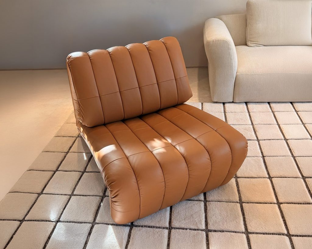 A low lounge armchair with generous channel tufting and warm tan leather upholstery offers a relaxed sculptural form. Its rounded silhouette and deep cushioned seat create an inviting balance of comfort and contemporary style.