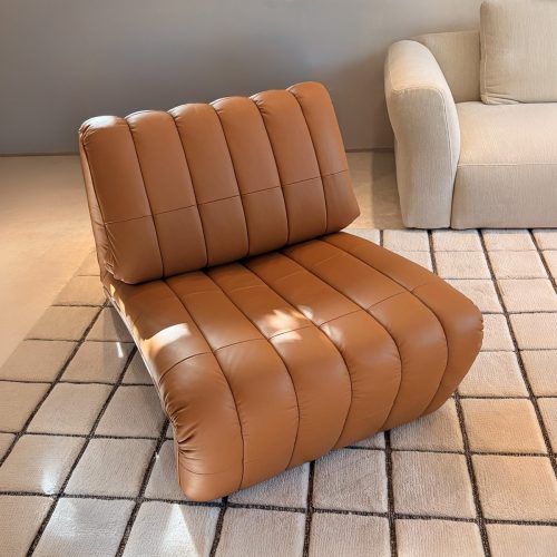 A low lounge armchair with generous channel tufting and warm tan leather upholstery offers a relaxed sculptural form. Its rounded silhouette and deep cushioned seat create an inviting balance of comfort and contemporary style.