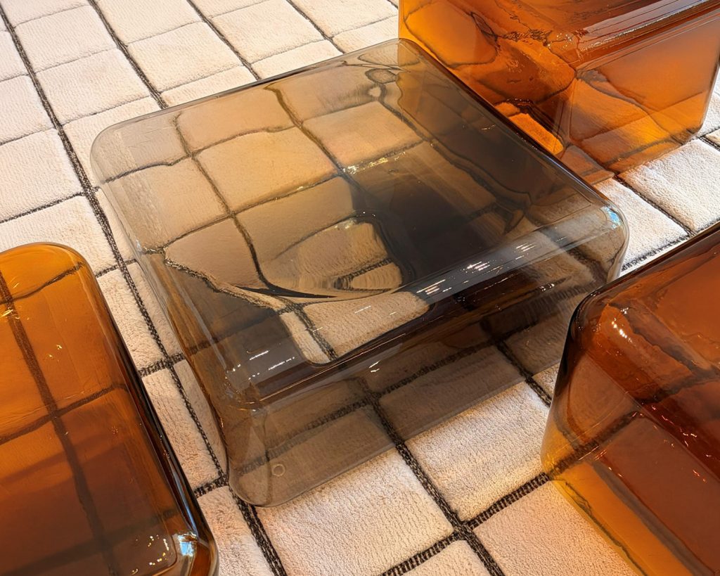 A low coffee table with softly rounded edges and a translucent smoky glass finish creates a light, sculptural presence. Its seamless form reflects surrounding light, bringing depth, warmth, and a refined contemporary character to the space.