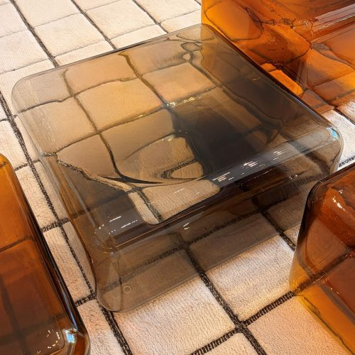A low coffee table with softly rounded edges and a translucent smoky glass finish creates a light, sculptural presence. Its seamless form reflects surrounding light, bringing depth, warmth, and a refined contemporary character to the space.